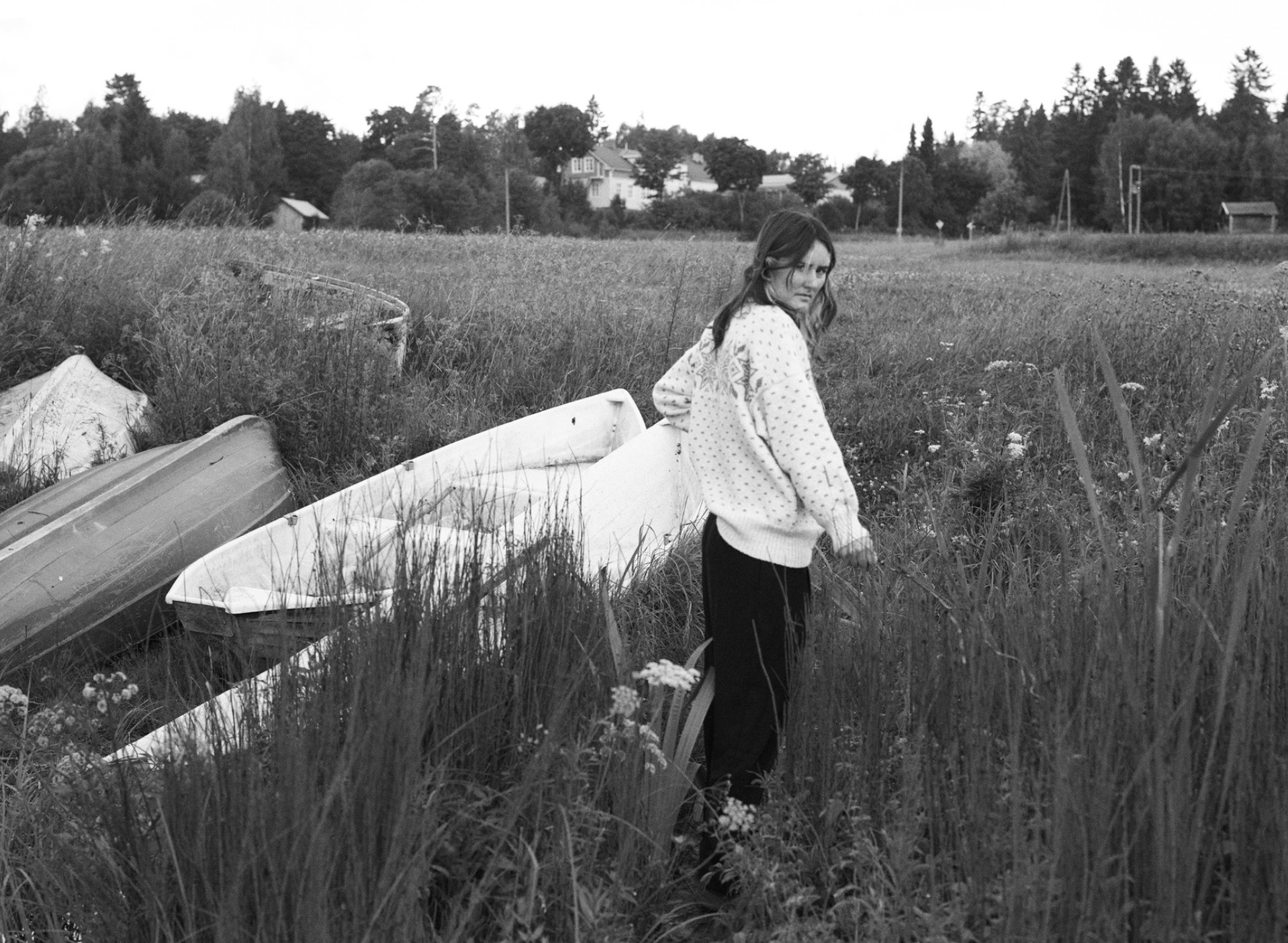 Figure in a knit sweater standing among overturned rowboats in a field