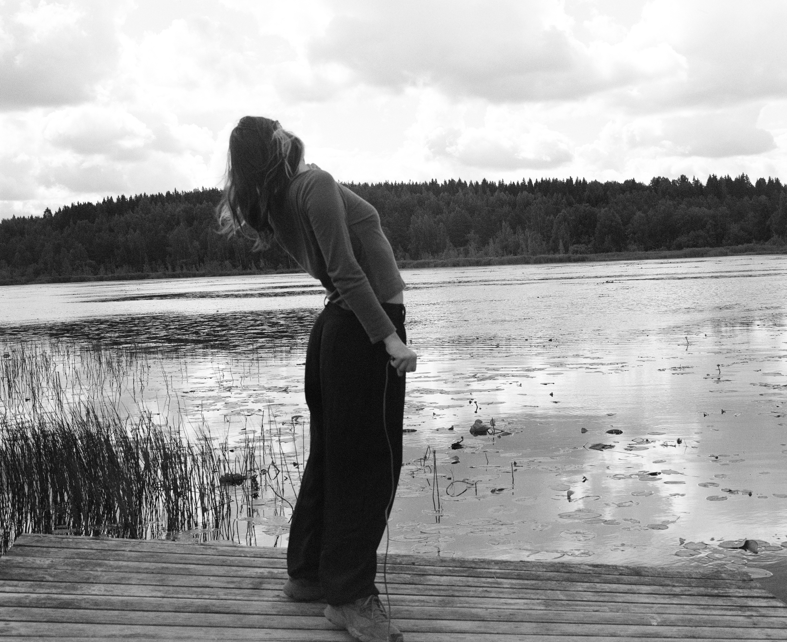 Figure bent forward on a wooden jetty, looking down at a lily-pad-covered lake