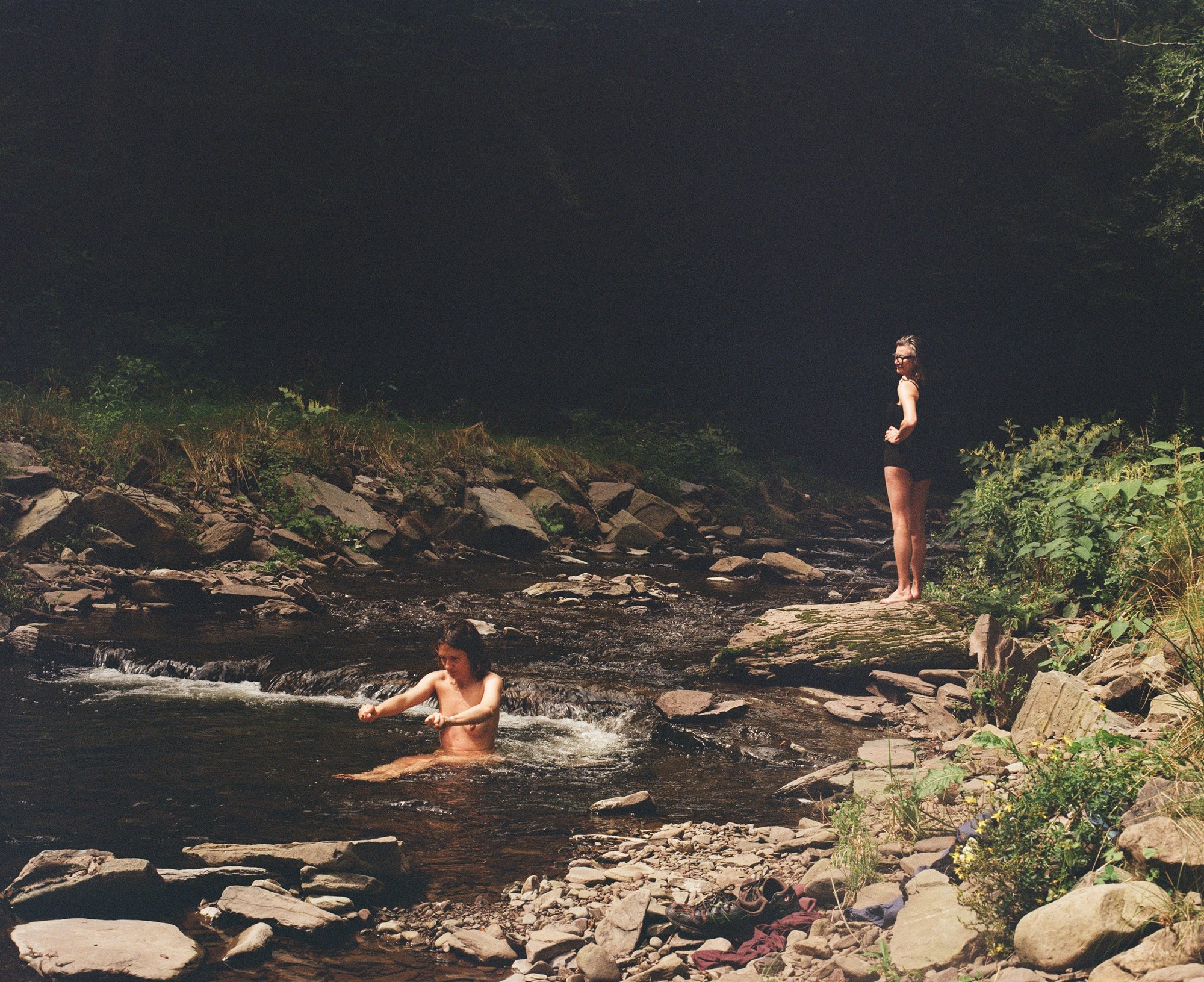 A person swimming in a creek while another stands watching on a rock, upstate New York