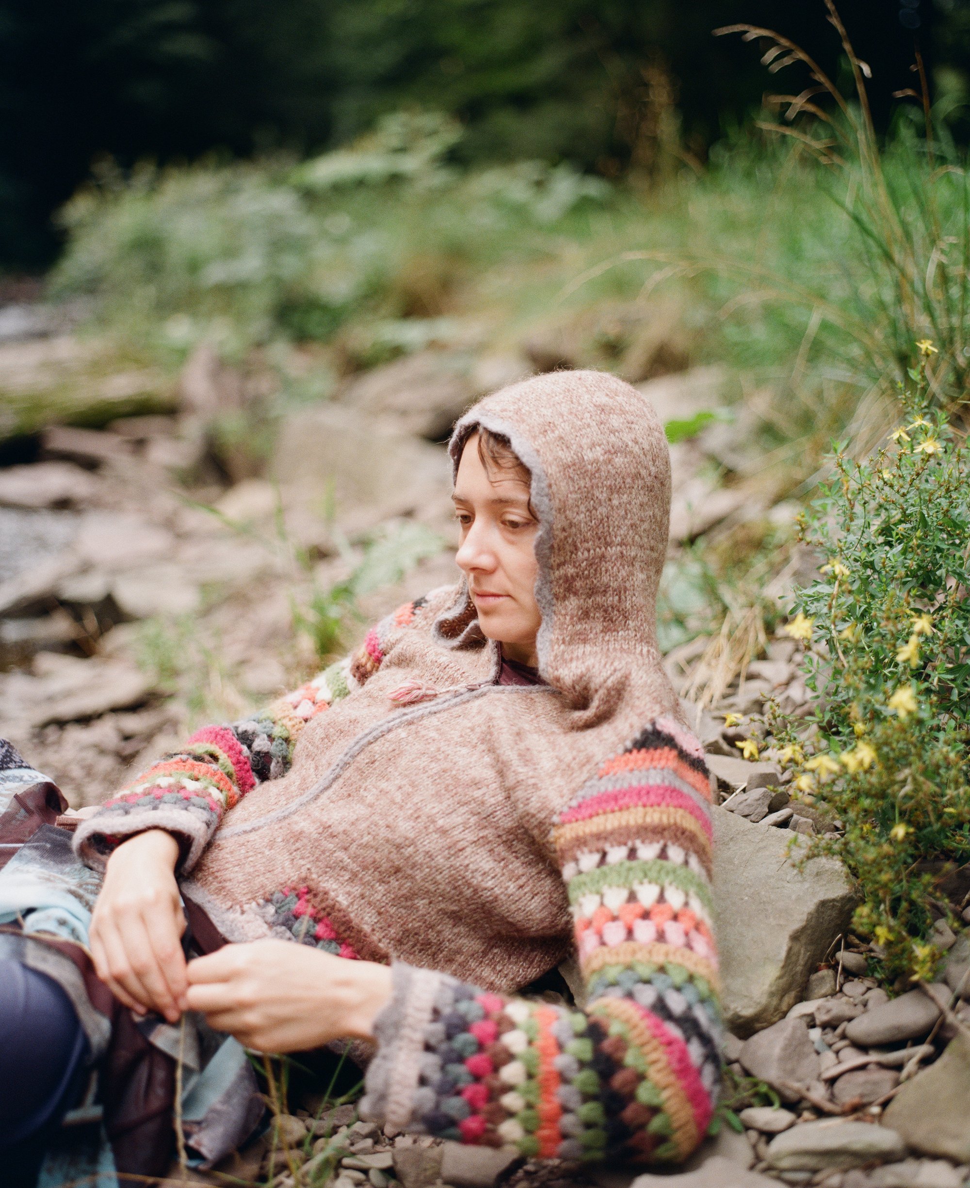 Woman in a striped knit hood and cardigan resting on rocks by a creek