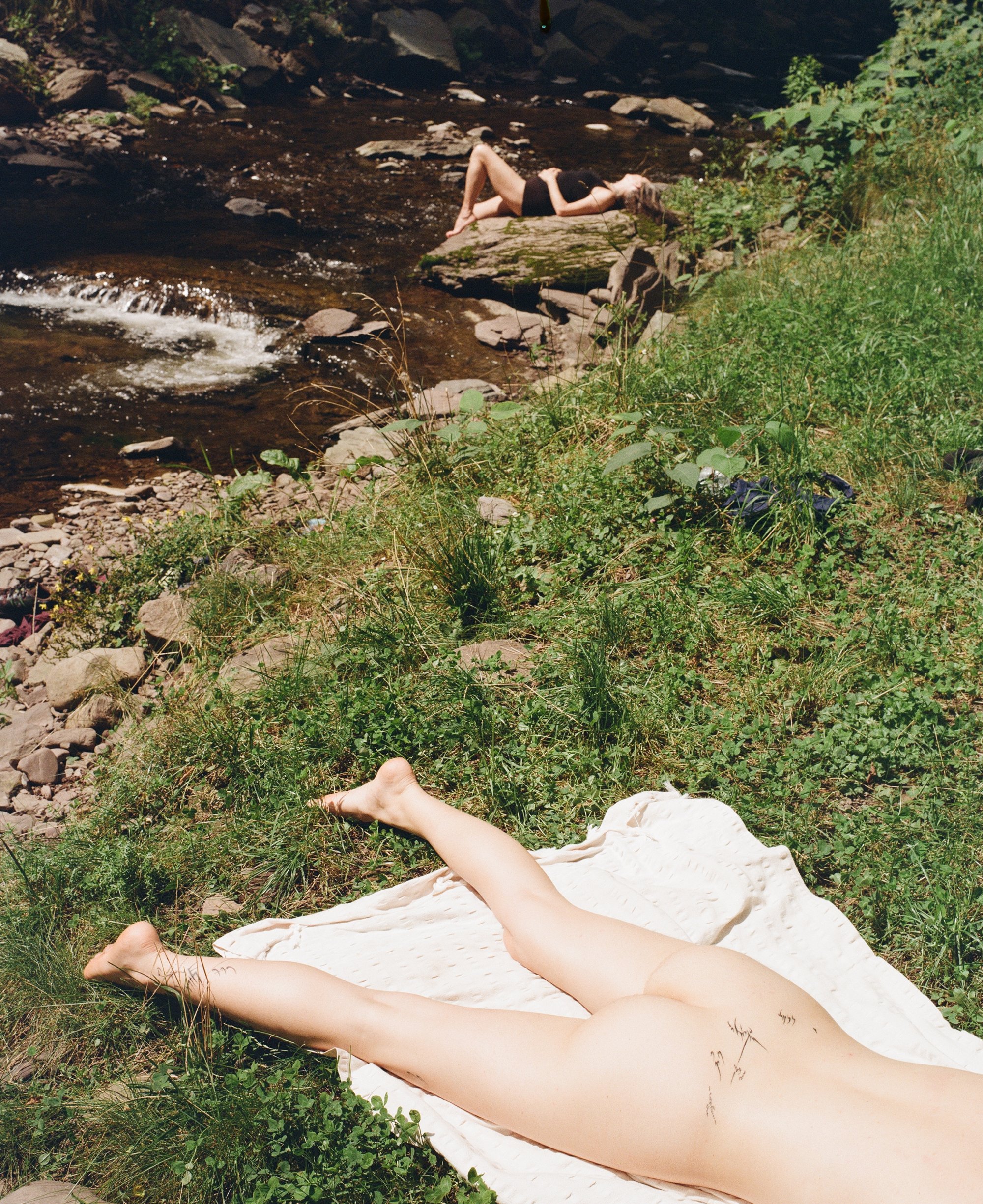 A person lying on a blanket by a creek, another sunbathing on rocks in the distance