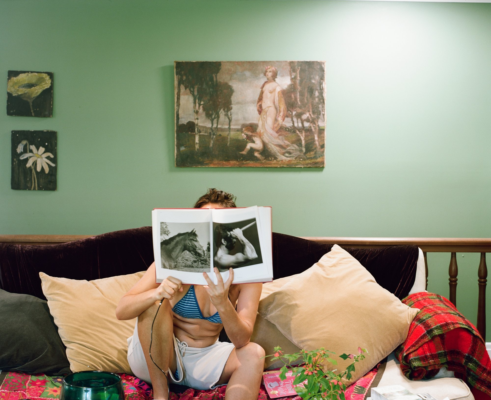 Woman in a bikini top holding an open book up over her face on a velvet sofa