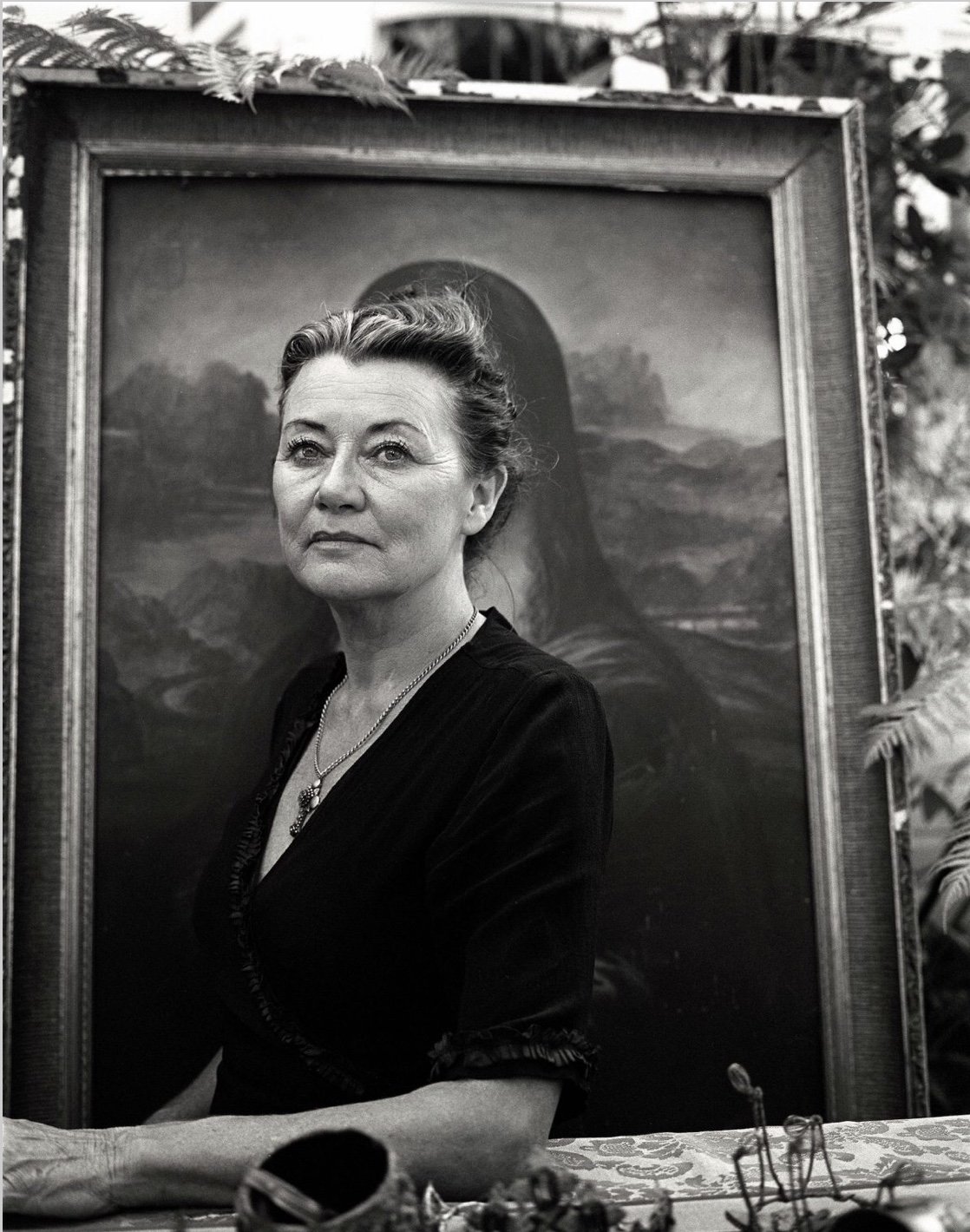 Portrait of an older woman in a black dress standing in front of a framed painting of the Mona Lisa
