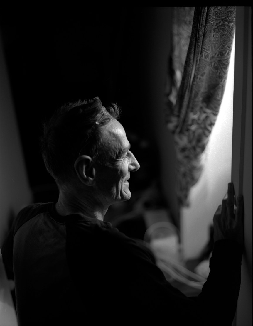 An older man in profile looking out toward a sunlit window, curtain drawn back