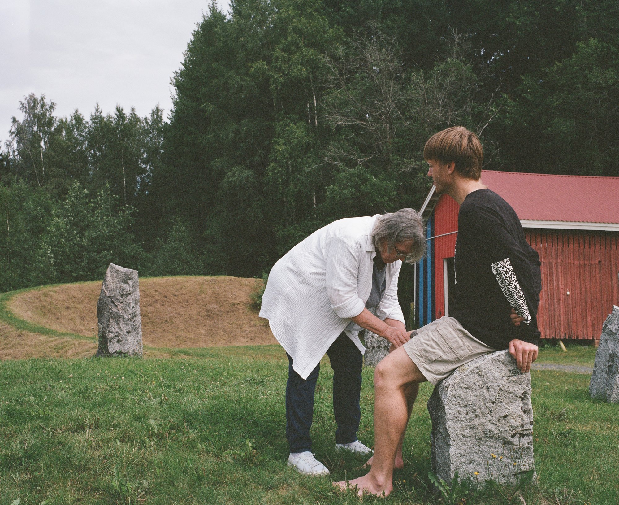 An older man bending to look at a younger man's leg, seated on a standing stone