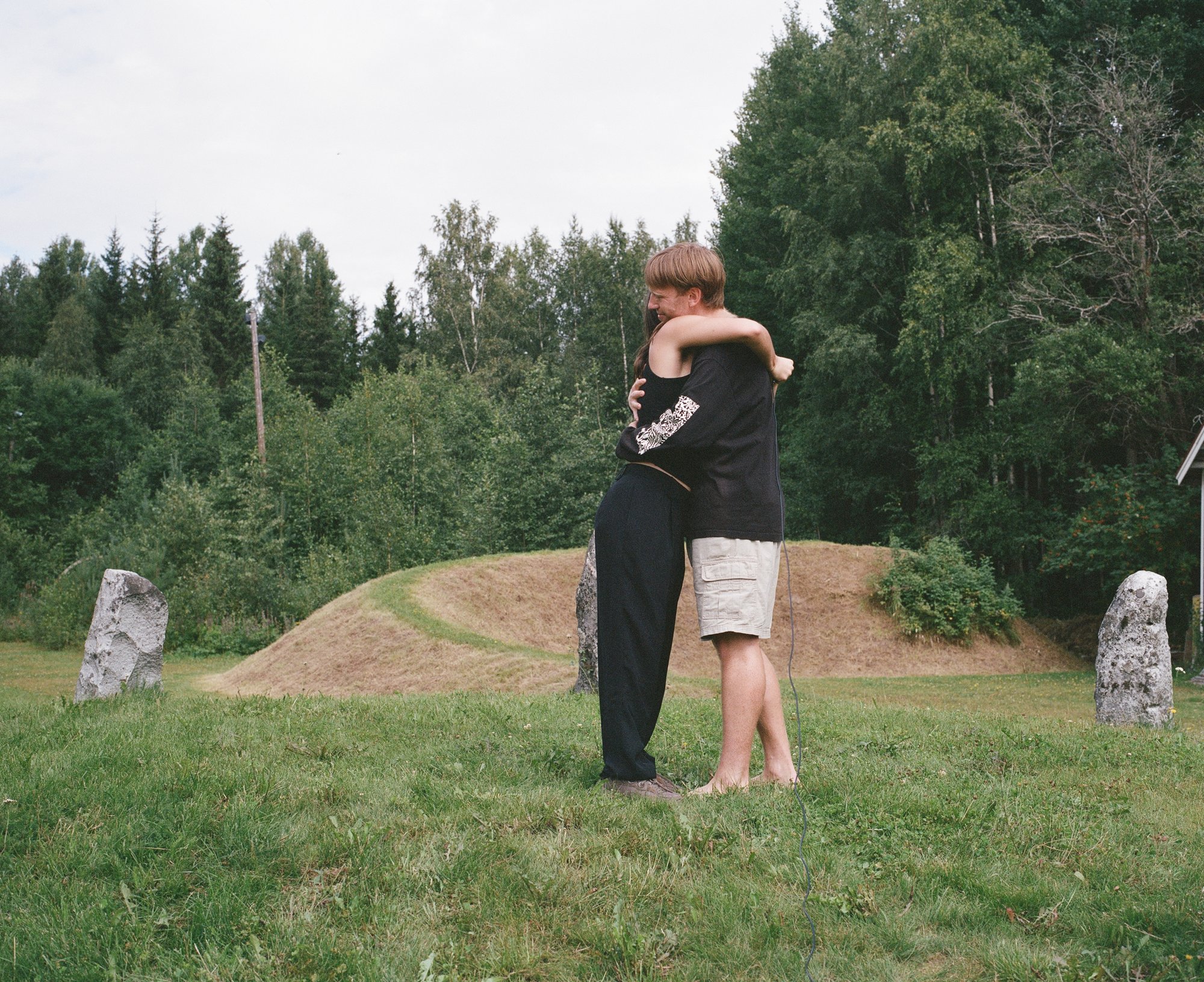 Two figures embracing among standing stones, an electrical cord trailing across the grass