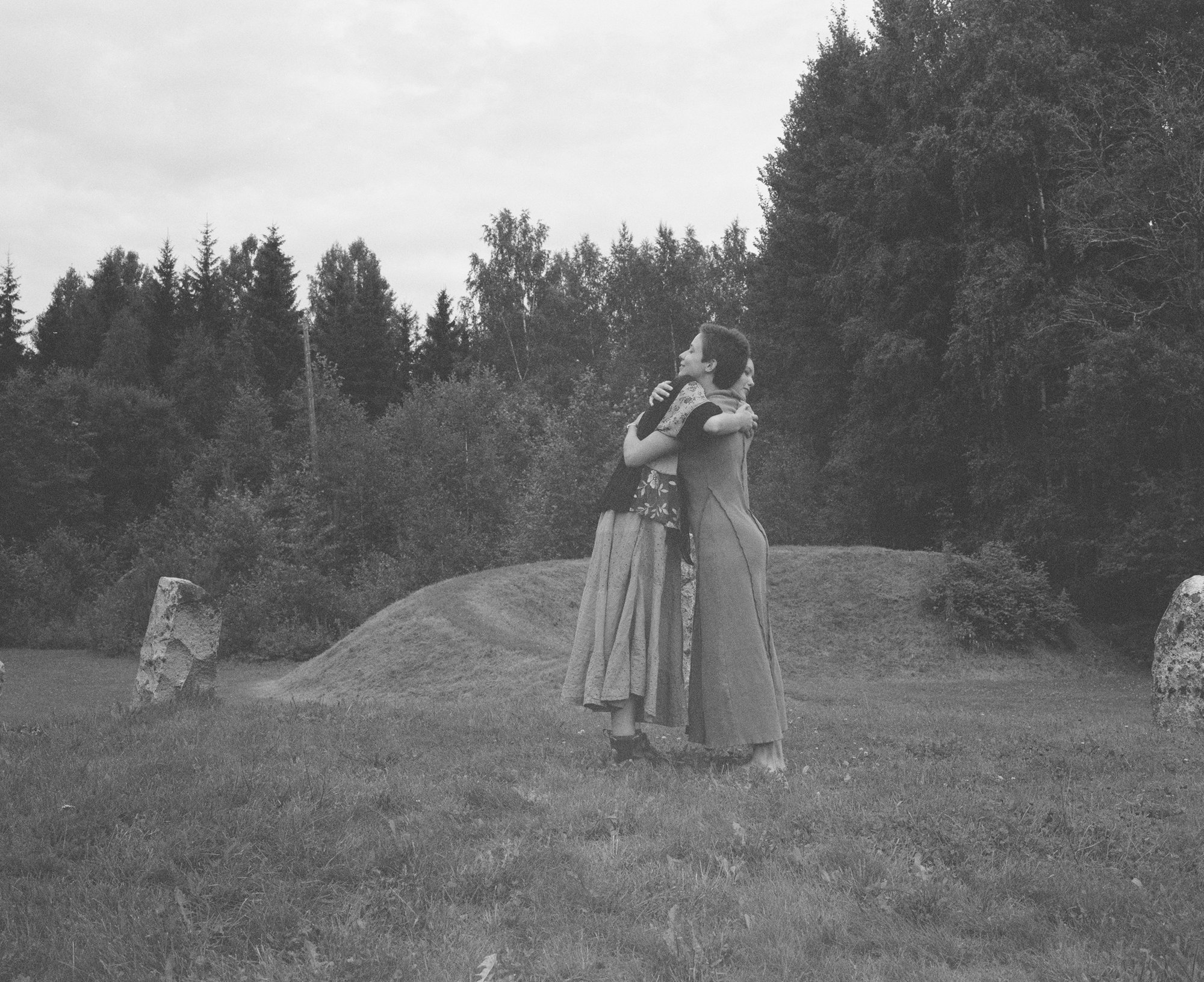 Black and white photograph of two figures in long skirts embracing among standing stones