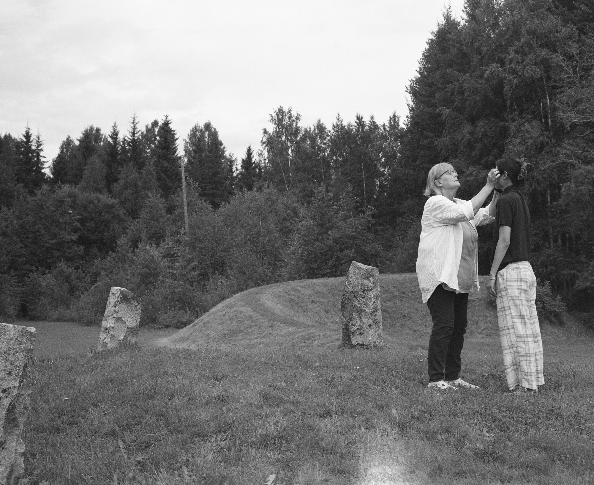Black and white photograph of an older woman adjusting another figure's face among standing stones
