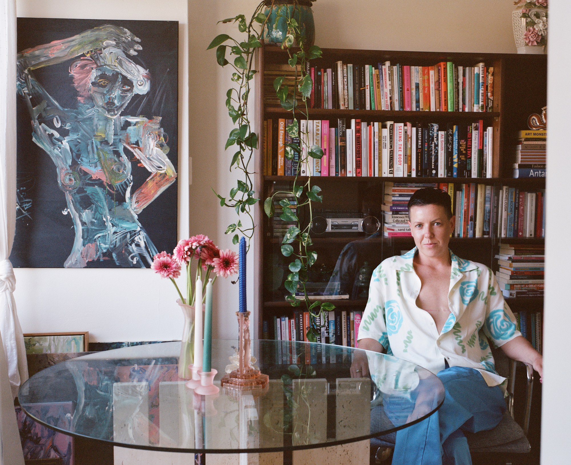 Portrait of a person seated at a glass table in front of a bookshelf and painting