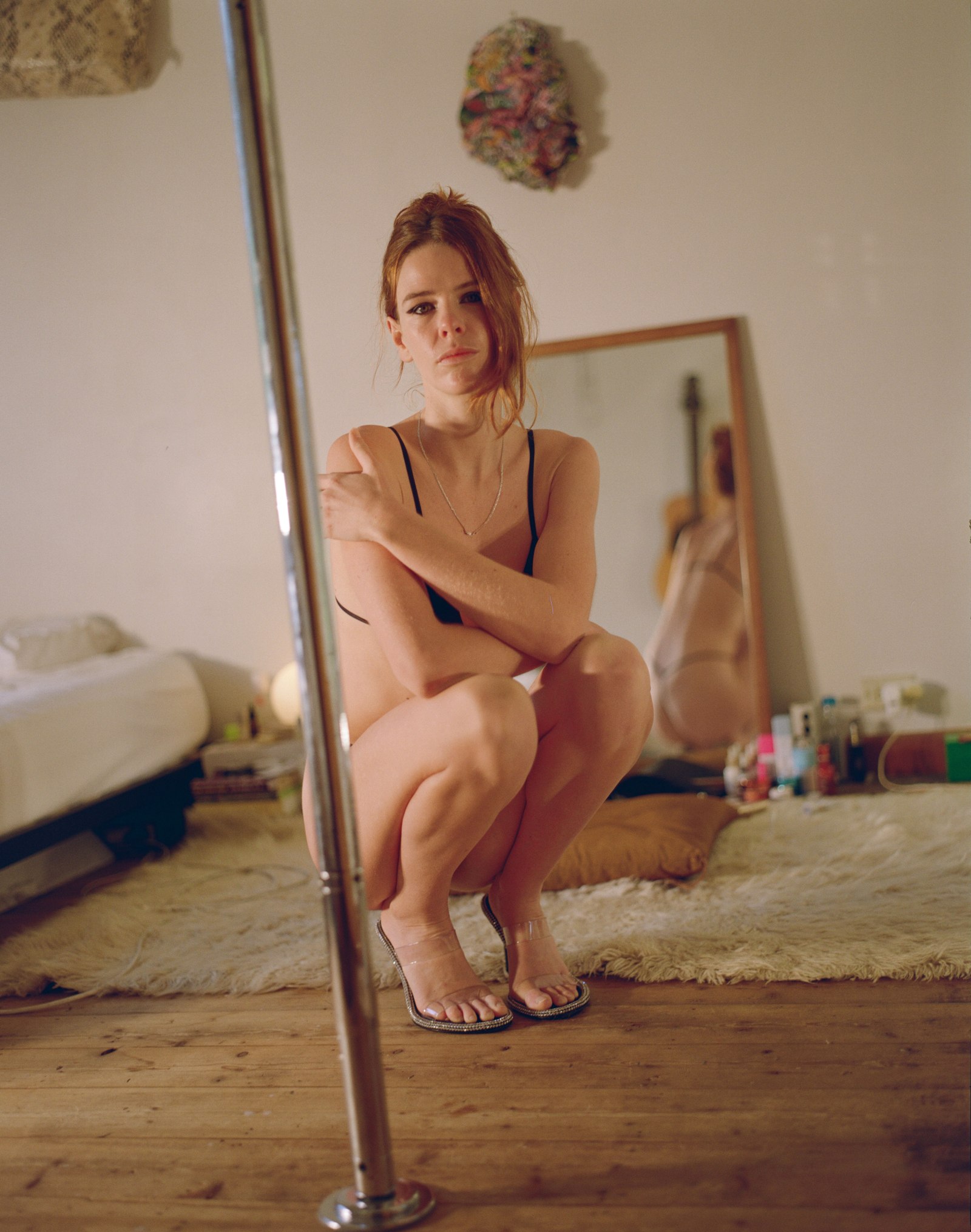 Portrait of a woman crouched beside a pole in a bedroom, a mirror reflecting her behind