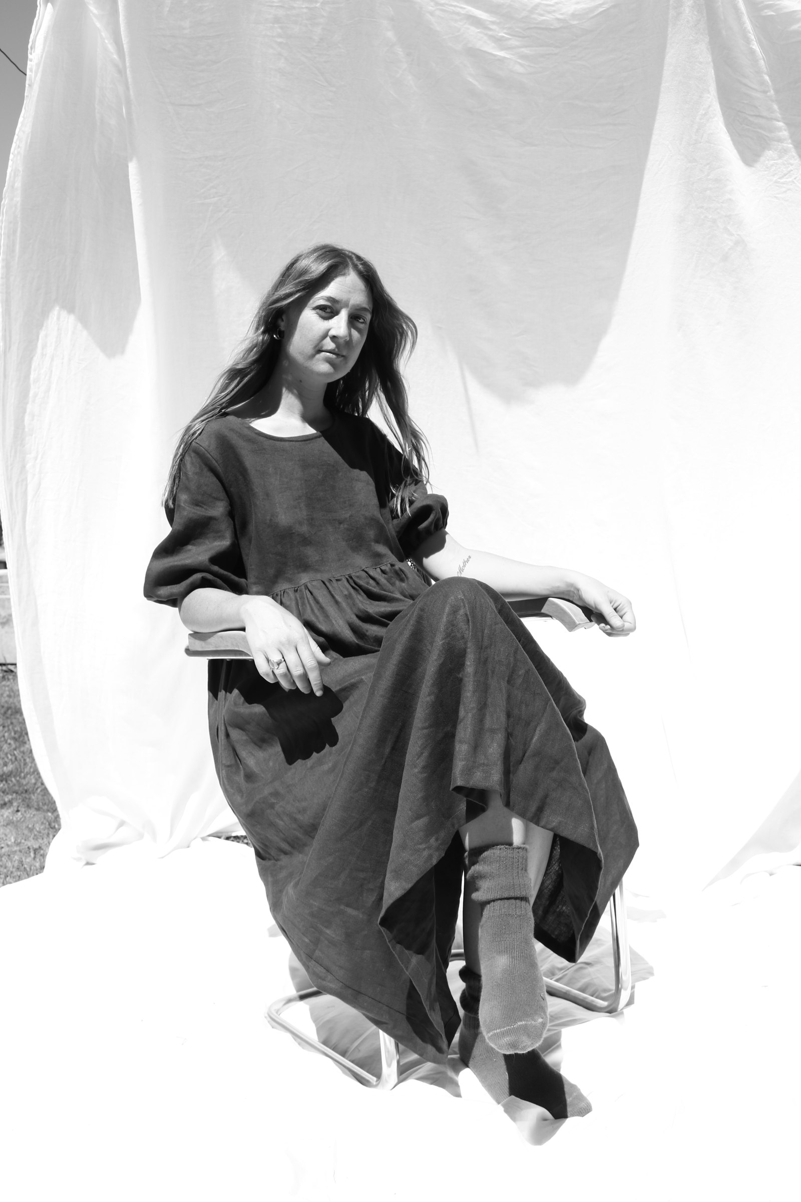 Portrait of a woman in a dark linen dress seated in front of a hanging white sheet
