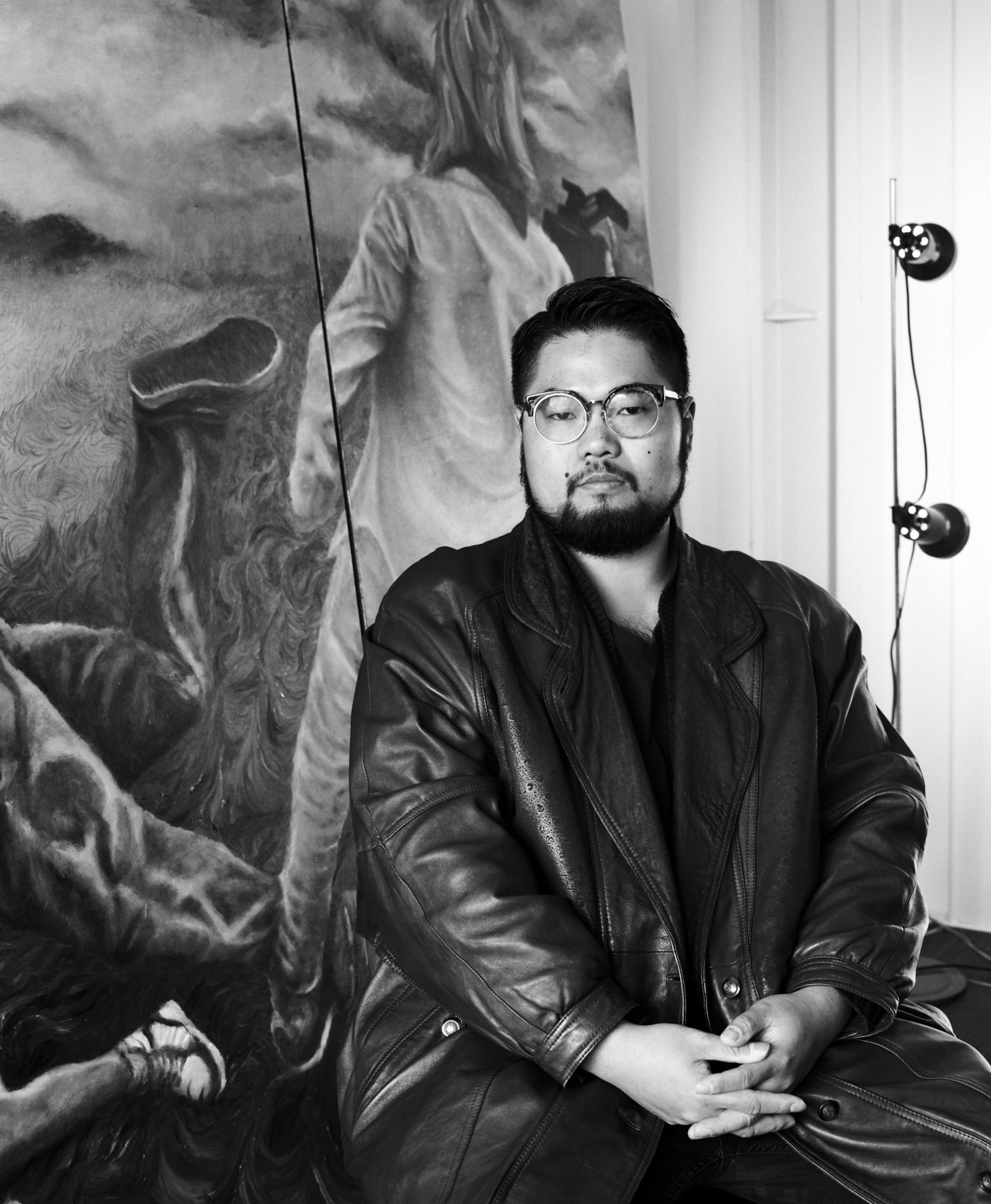 Portrait of a man in a leather jacket and glasses seated in front of a large charcoal painting