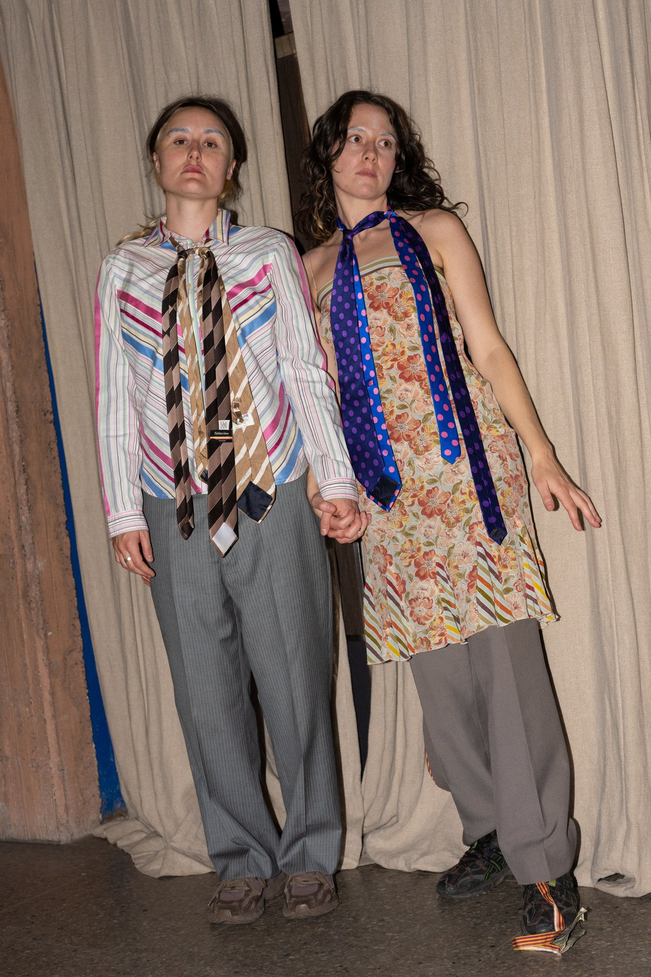 Two performers in grey suits with mismatched ties, one in a floral dress, standing against curtains