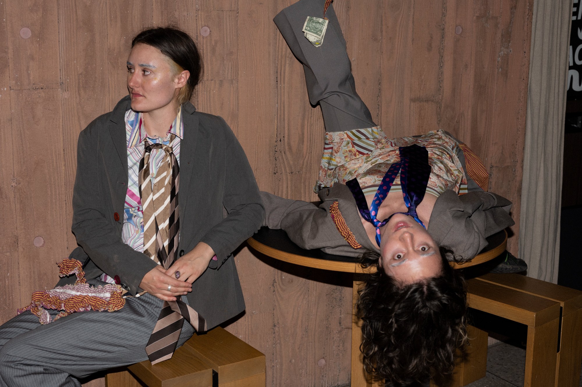 Two performers in a kitchen, one leaning over a table, one reaching for pots on a rack