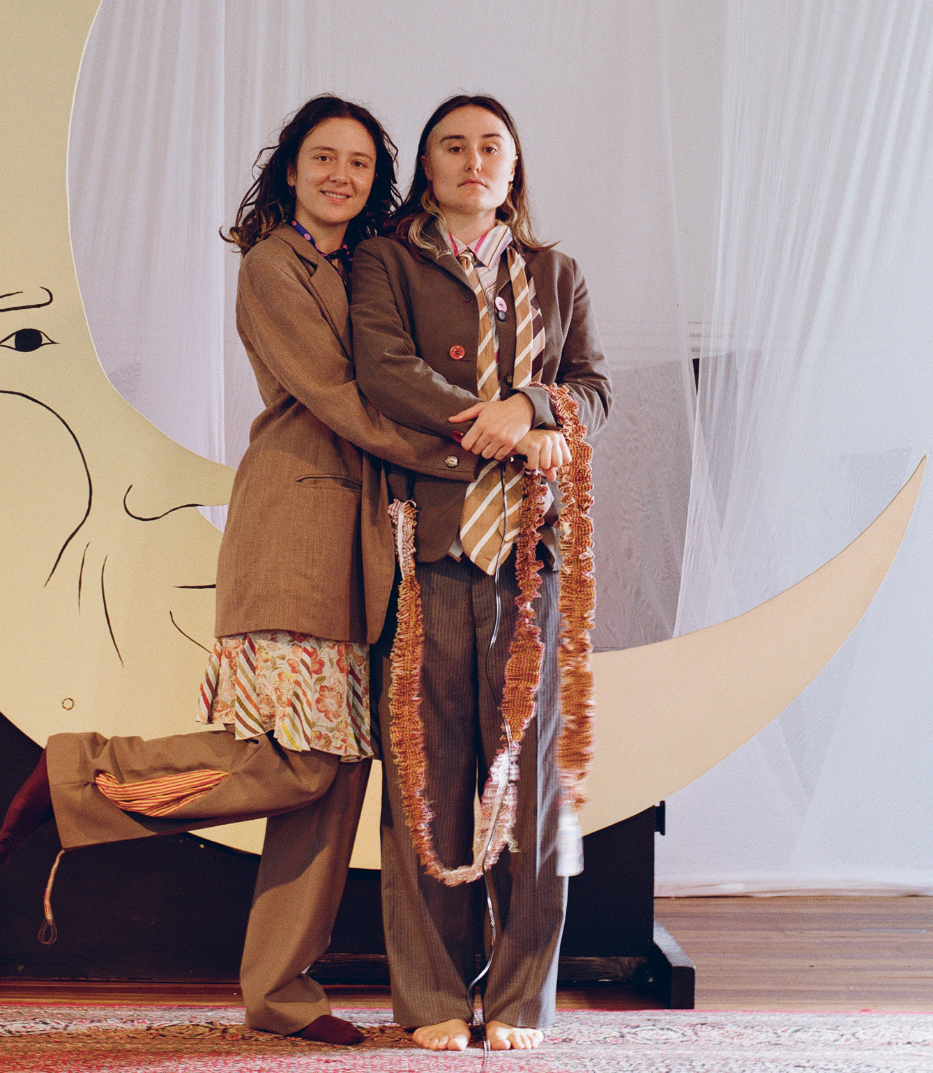 Two performers in matching brown suits embracing in front of a large crescent moon backdrop