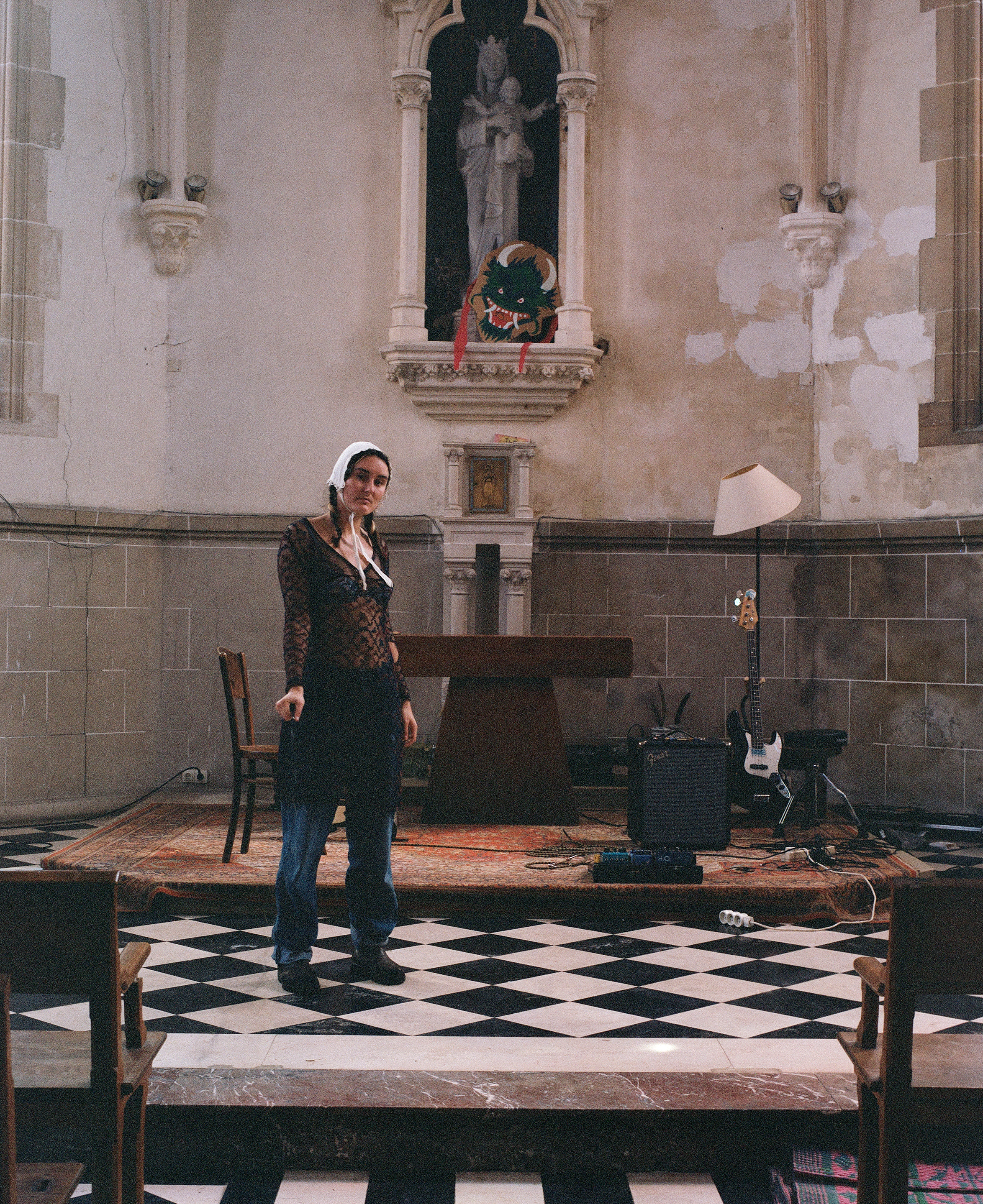 A performer in a lace top and headscarf standing on a checkered floor in a church, with band equipment set up behind her