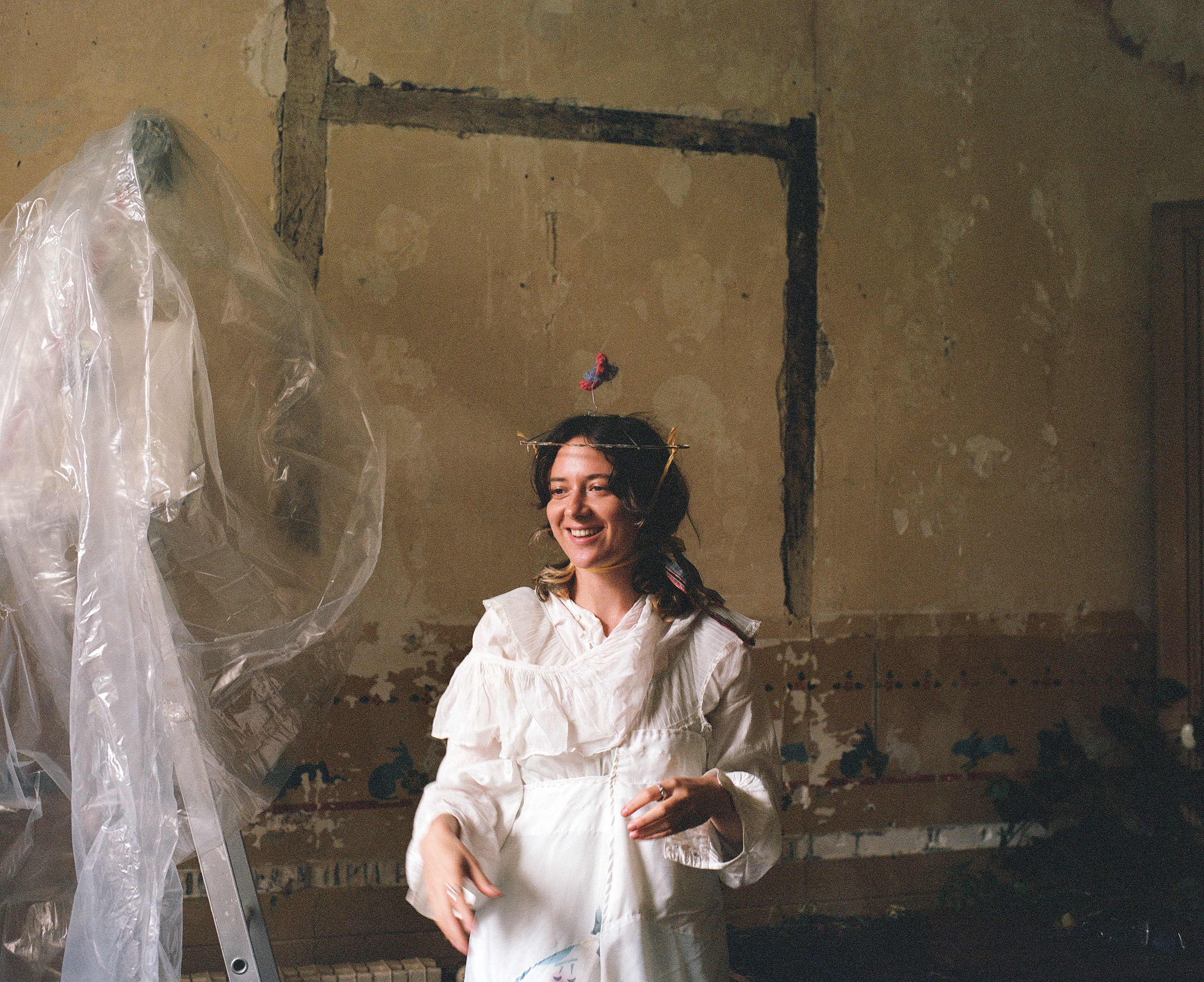 A performer in a white dress and twig crown, smiling, in front of a plastic-draped ladder in a derelict room