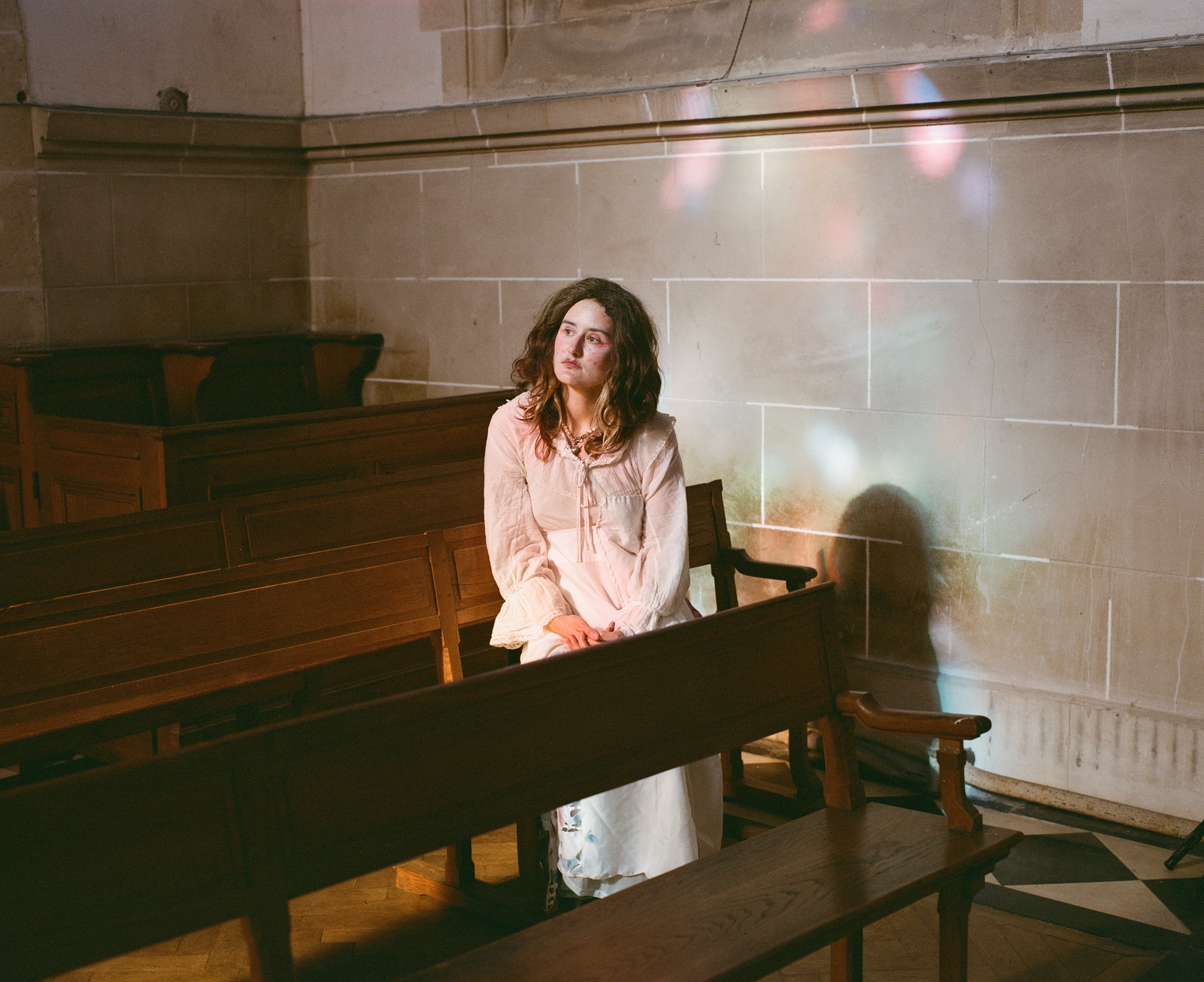 A performer in a white dress seated in a church pew, lit by colourful light through stained glass
