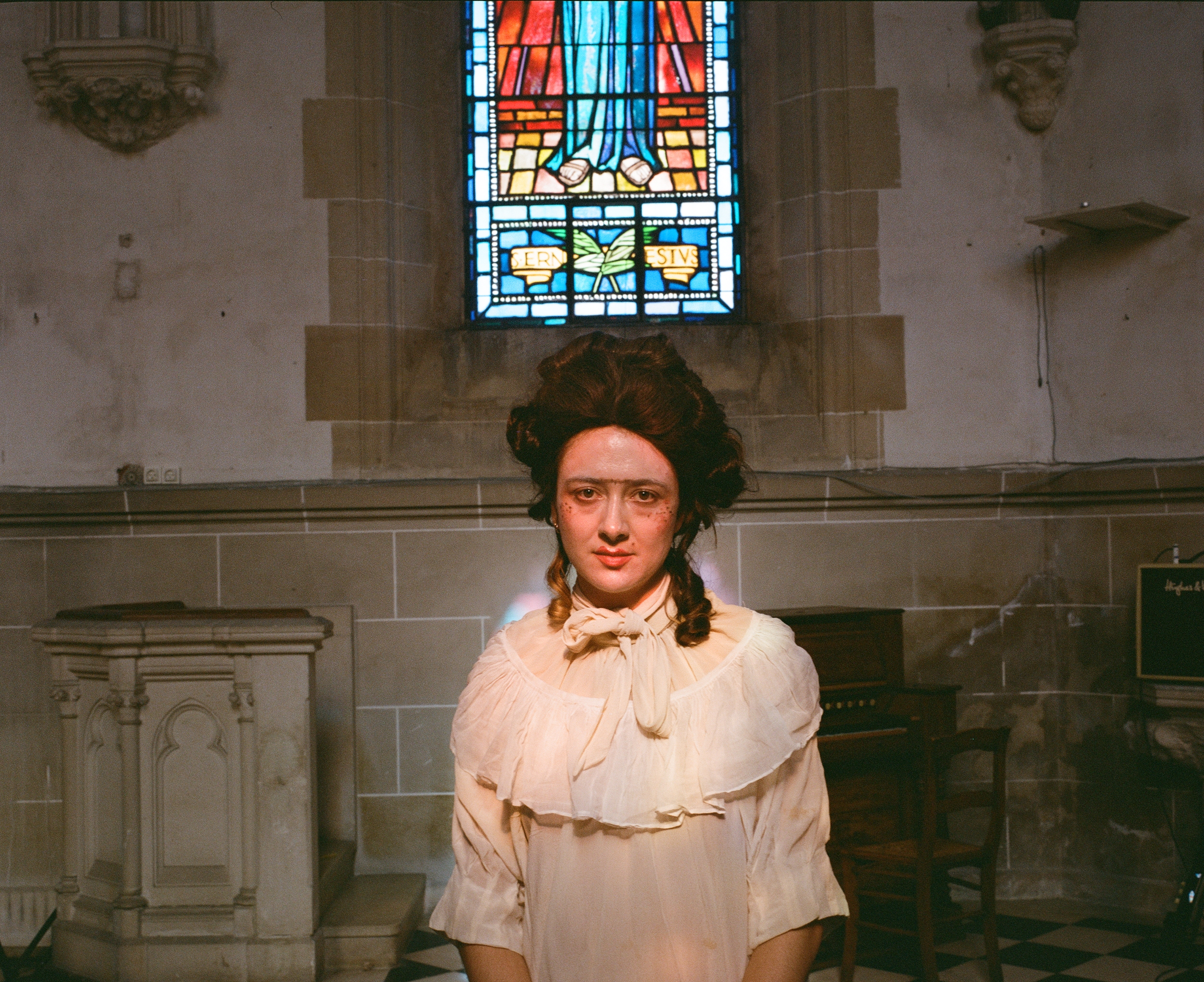 A performer in a powdered wig and cream blouse standing before a stained glass window