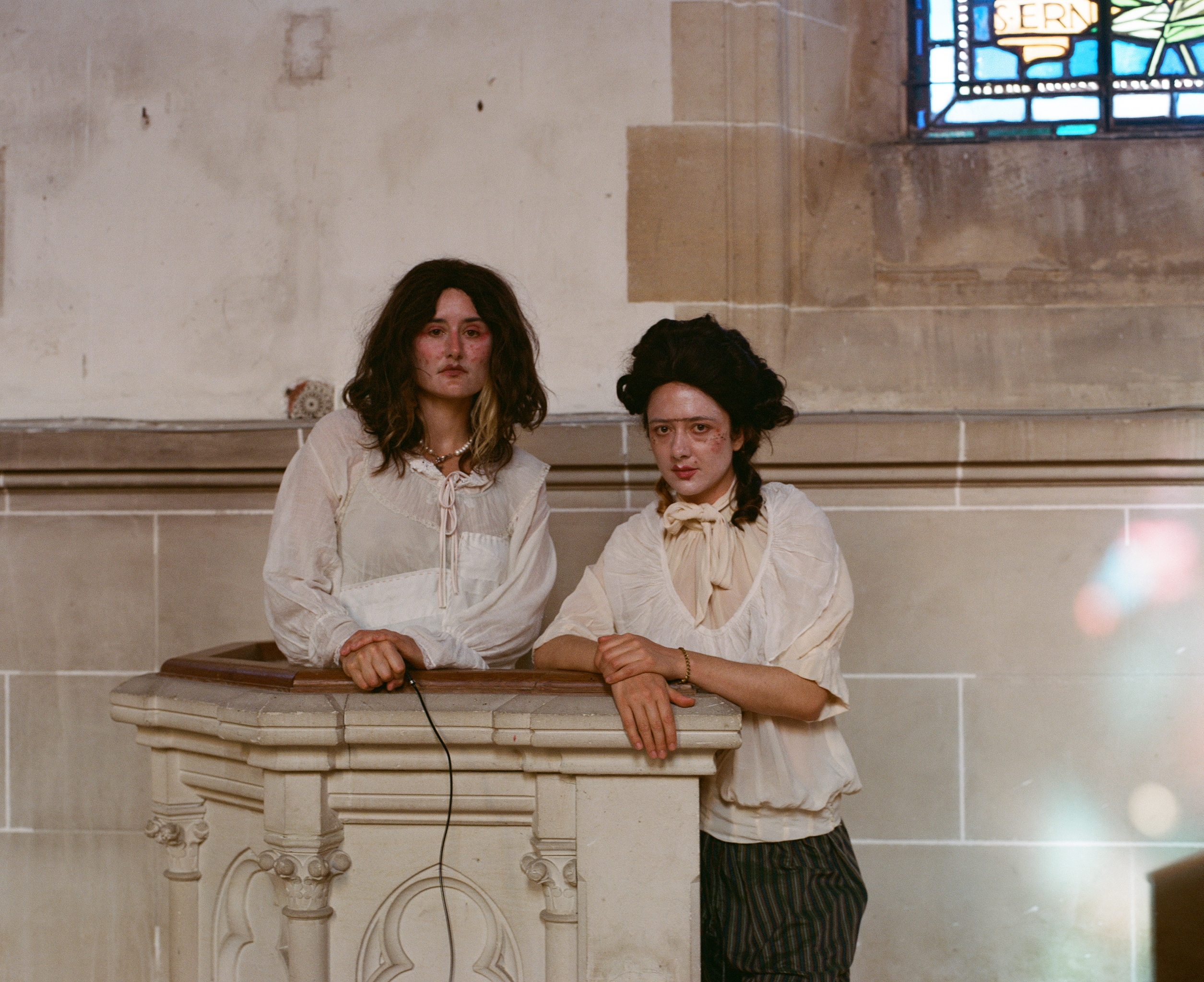 Two performers in wigs and white blouses leaning on a stone pulpit