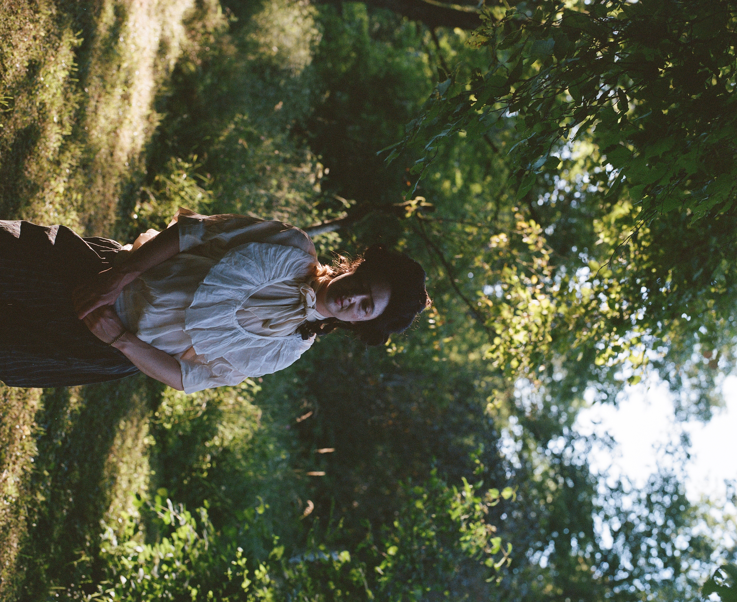 A performer in a powdered wig and ruffled blouse standing in dappled light in a garden