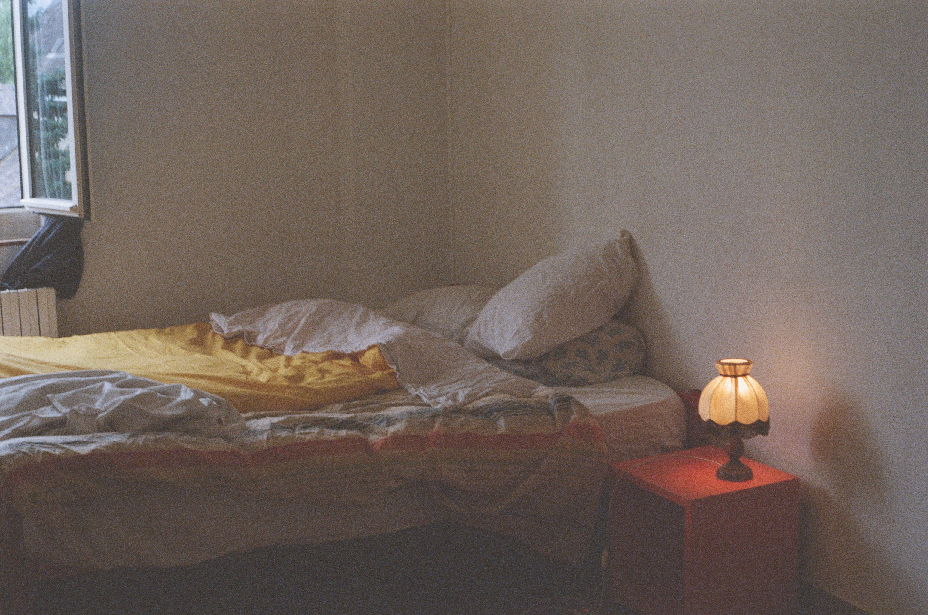 An unmade bed lit by a small lamp on a red bedside table, curtain blowing at an open window
