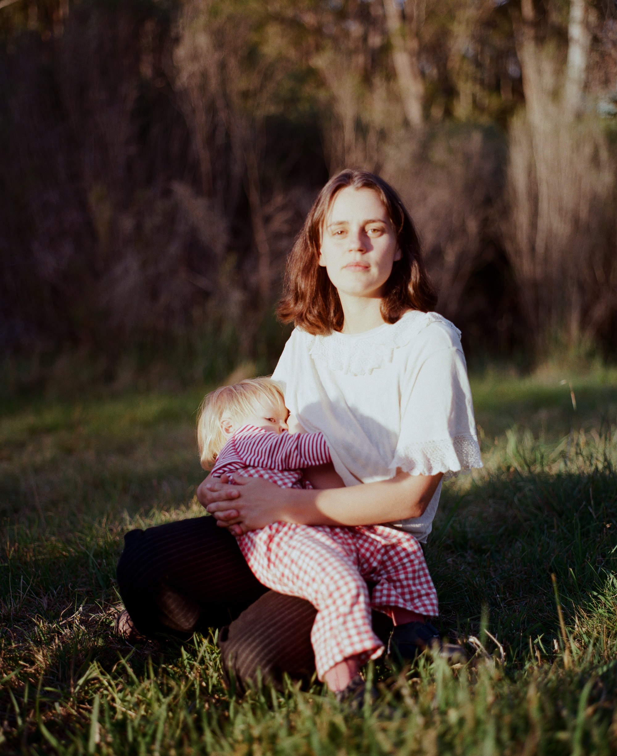 A mother breastfeeding her toddler while seated in a sunlit field